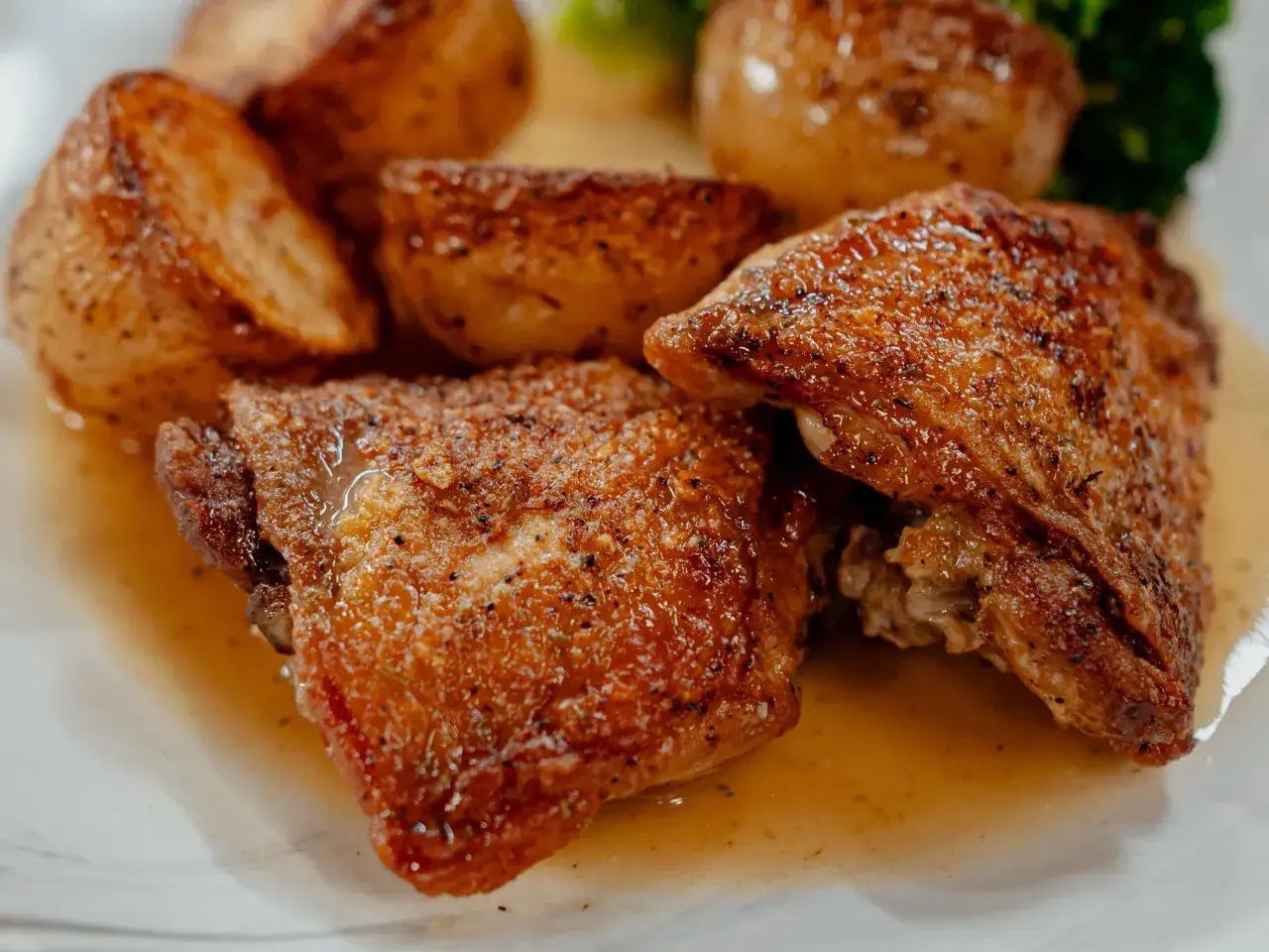 Deux morceaux de haut de cuisse de poulet au four, dorés et juteux, baignant dans leur jus, accompagnés de pommes de terre rôties et d'un peu de brocolis.