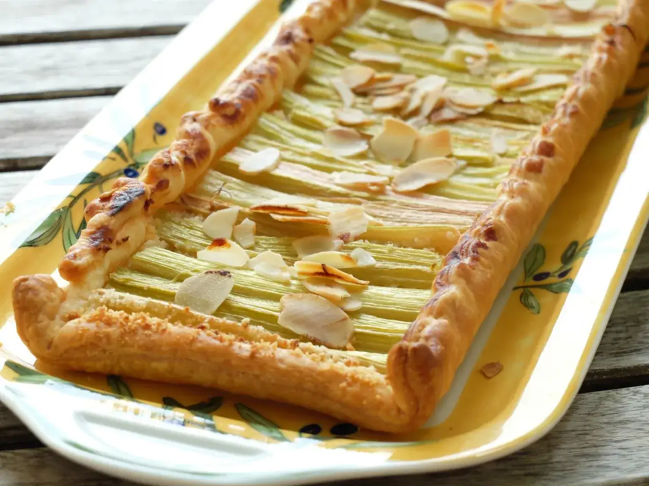 Tarte à la rhubarbe sur pâte feuilletée, garnie d'amandes effilées, sur un plat jaune décoré. Un délice croustillant et fruité.