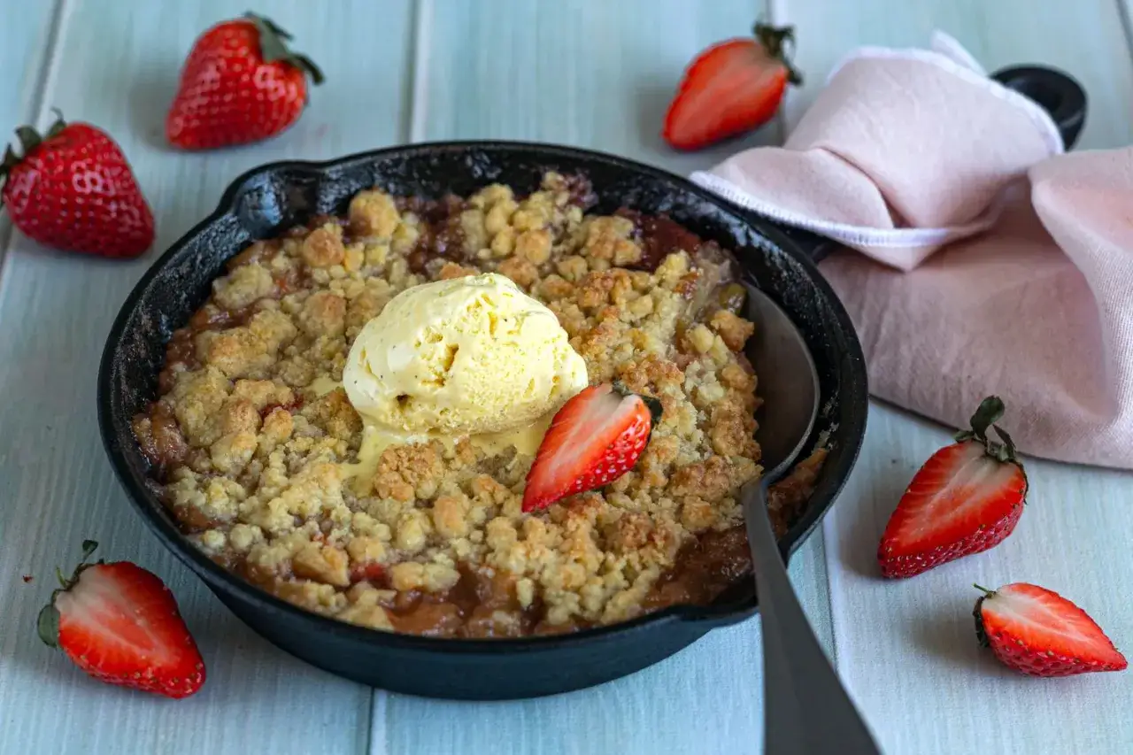 Un délicieux crumble rhubarbe fraise, servi chaud dans une poêle en fonte, surmonté d'une boule de glace à la vanille et décoré de fraises fraîches.