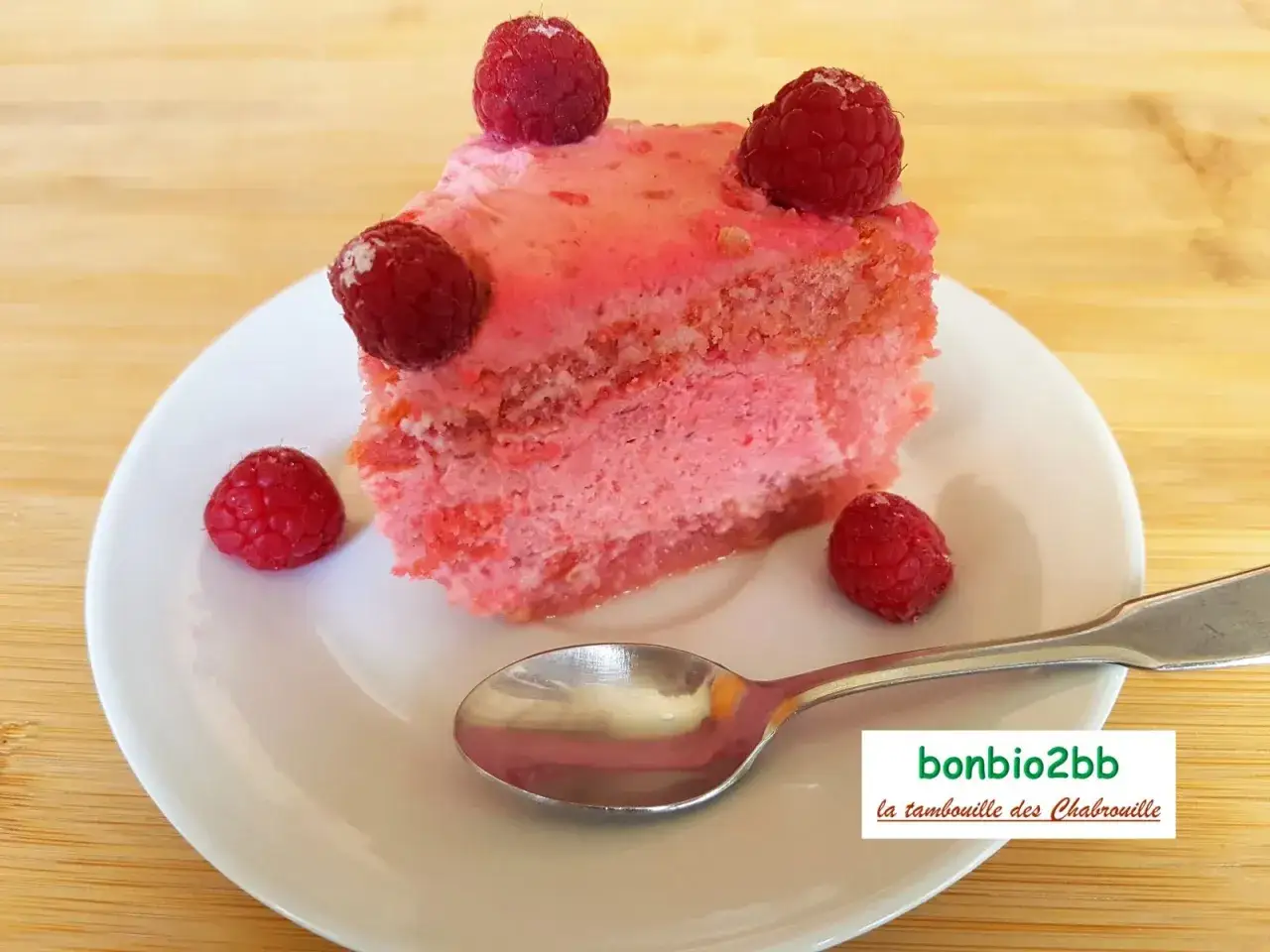 Une part de charlotte aux framboises, garnie de fruits rouges frais, sur une assiette blanche avec une cuillère.