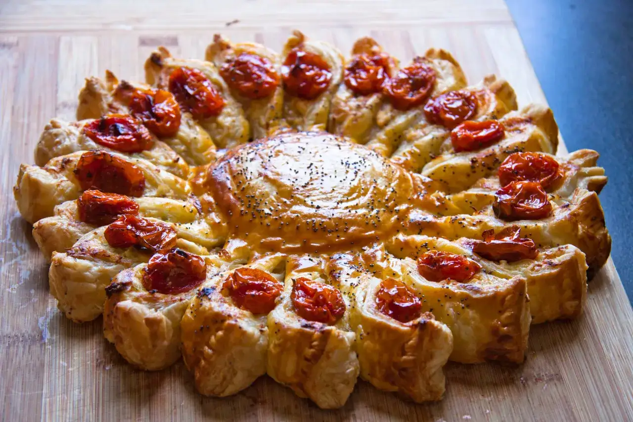 Tarte soleil dorée, garnie de tomates cerises confites et de graines de pavot, sur une planche en bois.