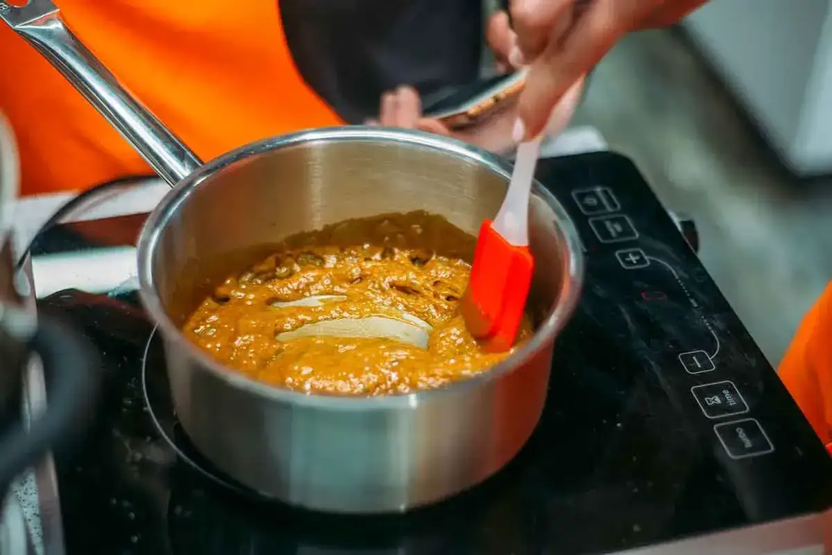 Une spatule rouge remue un caramel à sec bouillonnant dans une casserole en argent sur une plaque à induction.
