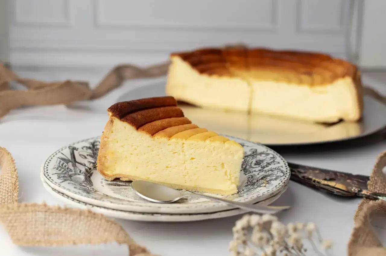 Une part de tarte au fromage blanc, légère et aérienne, sur une assiette ornée. Le reste du gâteau est visible en arrière-plan.
