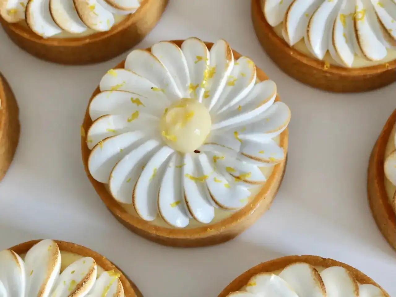 Plusieurs petites tartelettes au citron meringuées, garnies de crème jaune et de meringue blanche légèrement dorée, saupoudrées de zestes de citron.