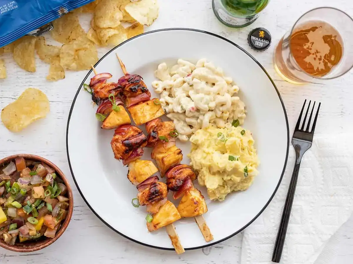 Brochettes de poulet grillé avec ananas et oignons rouges, accompagnées de salade de macaronis et de purée de pommes de terre. Un repas parfait pour un barbecue.