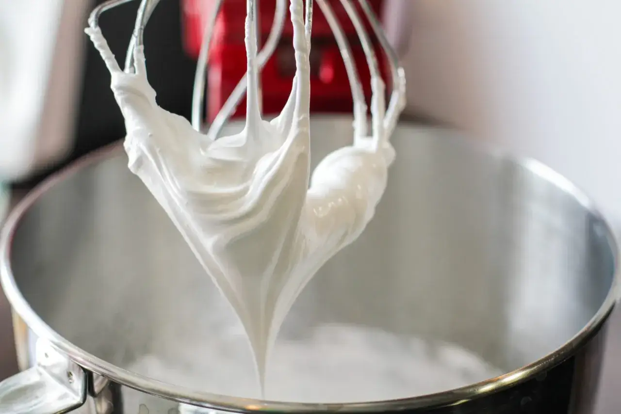 Meringue d'une blancheur en neige, tombant des fouets d'un robot pâtissier dans son bol en métal.