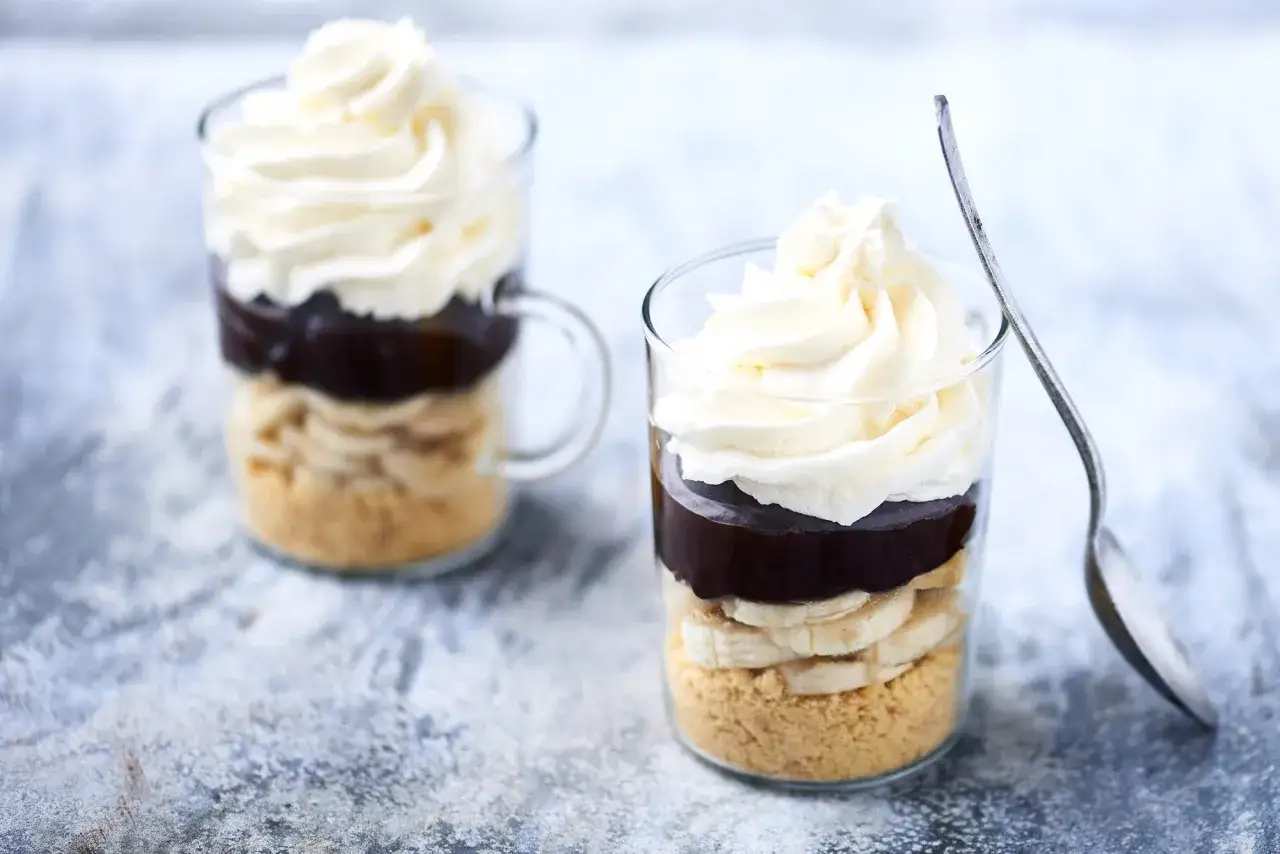 Deux délicieux banana split en verrine, superposant biscuits émiettés, rondelles de banane, sauce chocolat et crème fouettée.