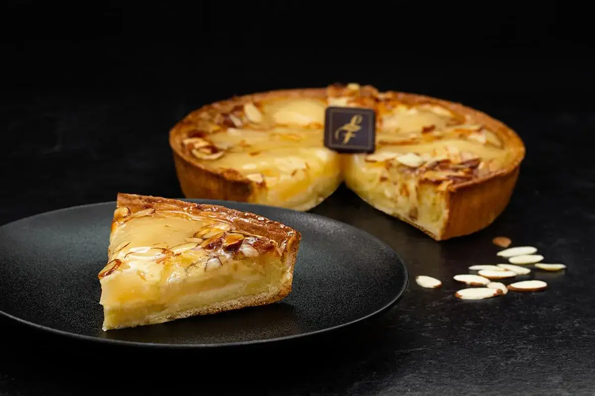 Une part de tarte aux poires, garnie d'amandes effilées et d'un glaçage brillant, est posée sur une assiette noire. Une tarte entière, décorée d'un logo, est visible en arrière-plan.