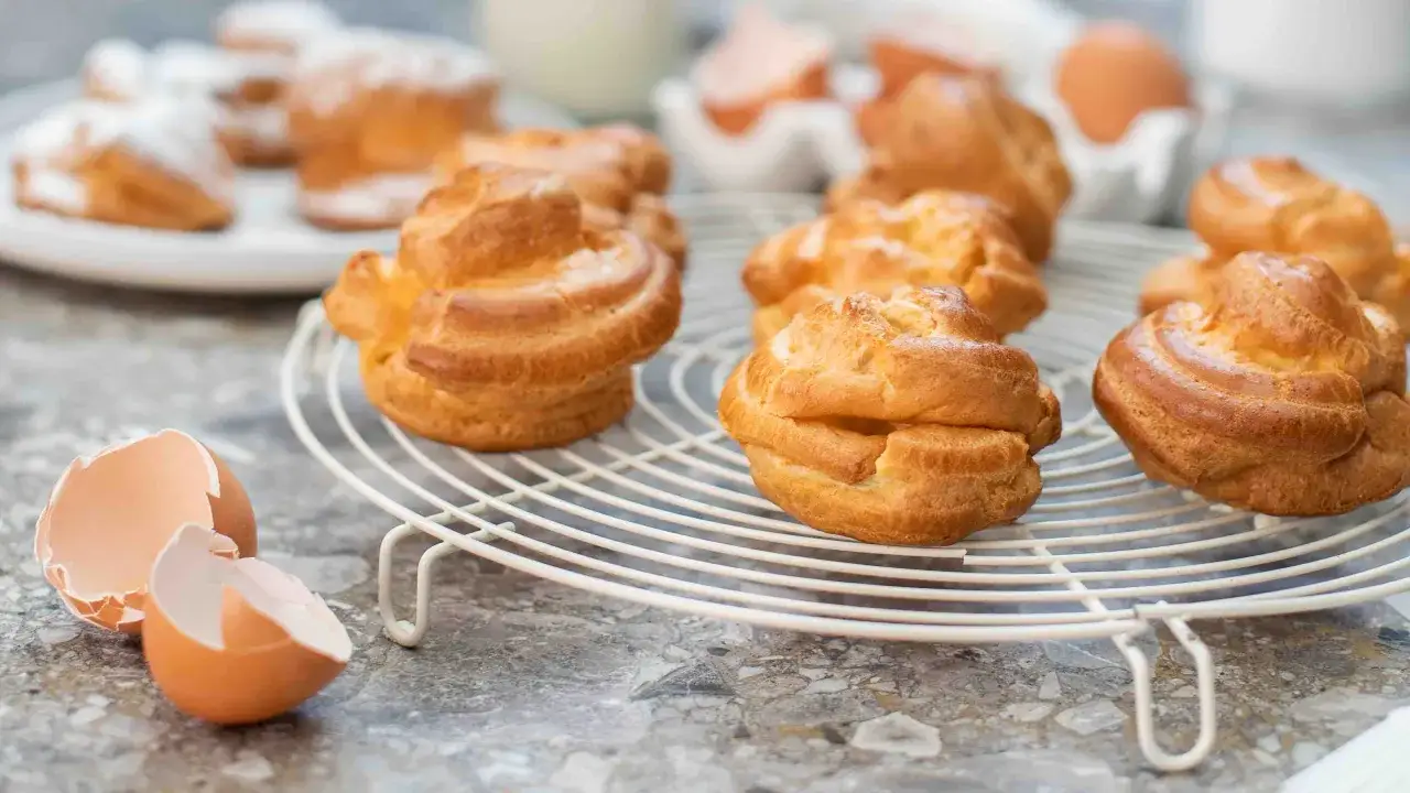 Des éclairs dorés de pâte à choux refroidissent sur une grille. Des coquilles d'œufs vides sont à proximité, rappelant les ingrédients de cette gourmandise.