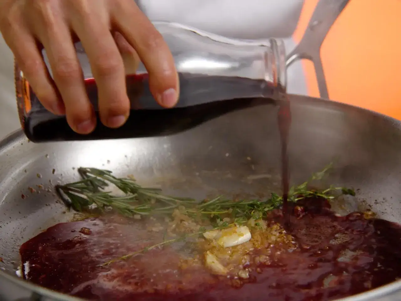 Une main verse un liquide rouge foncé dans une poêle contenant de l'ail, des herbes et du jus de viande.