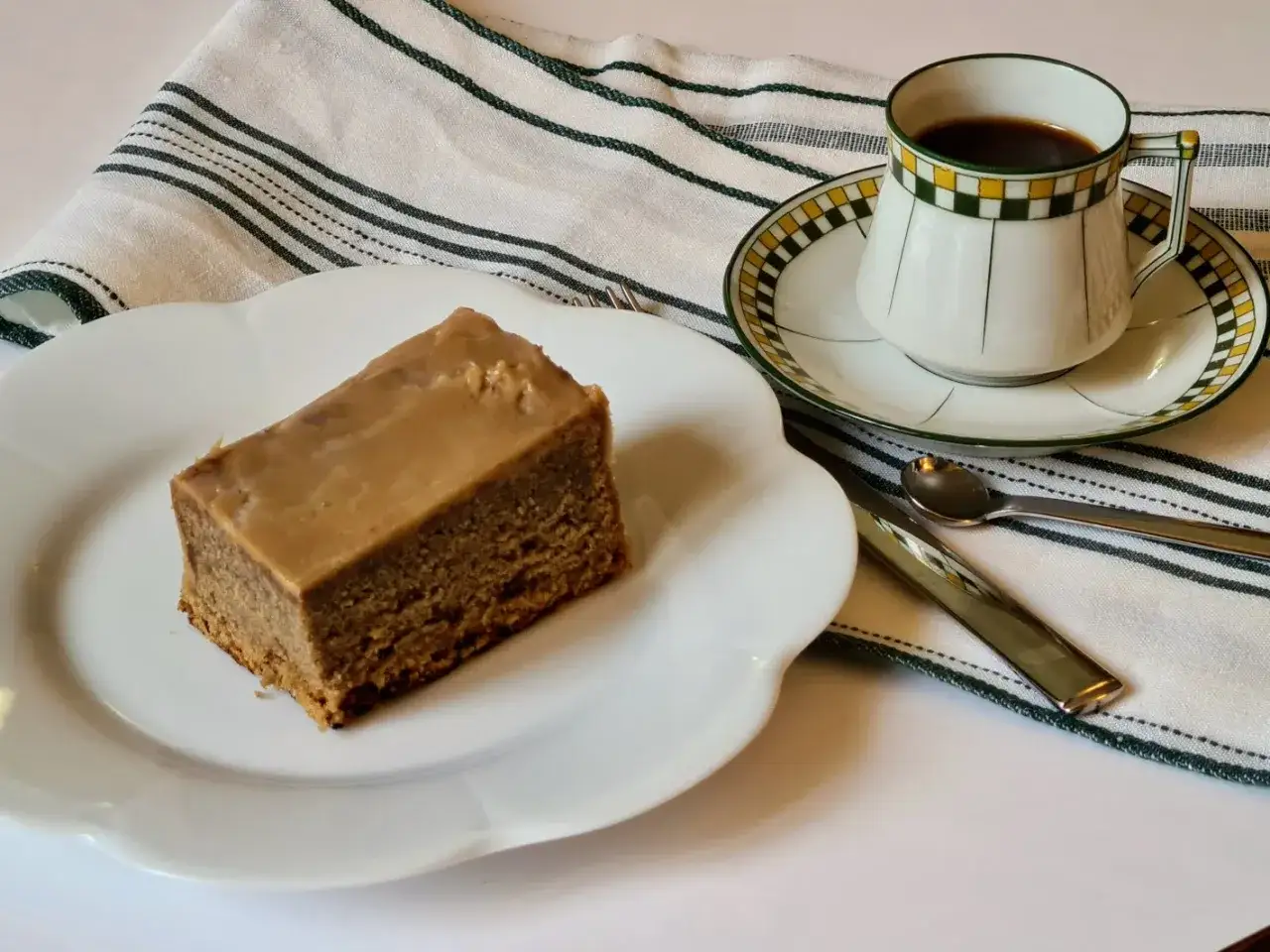 Tranche de gâteau au café glacé sur une assiette blanche, avec une tasse de café noir et des couverts sur une serviette rayée.