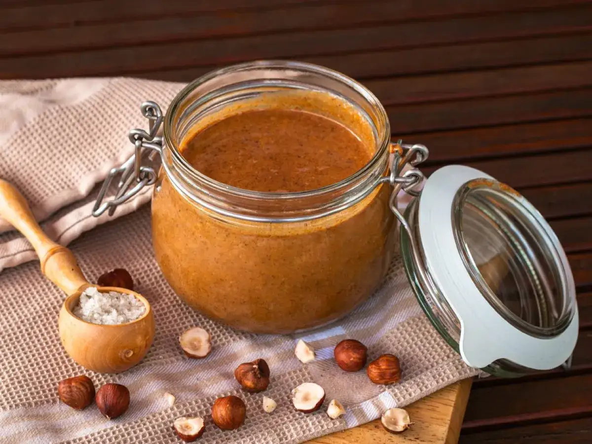 Un pot de praliné noisette maison, avec des noisettes entières et concassées autour. Une cuillère en bois contient du sel, suggérant une recette gourmande.