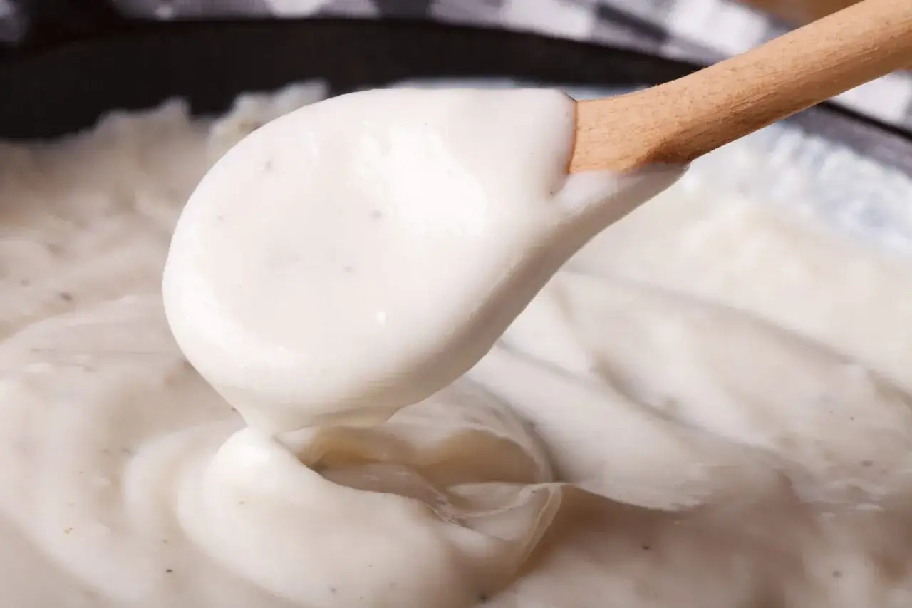 Une cuillère en bois prélève une généreuse portion de sauce blanche onctueuse, prête à napper un plat.