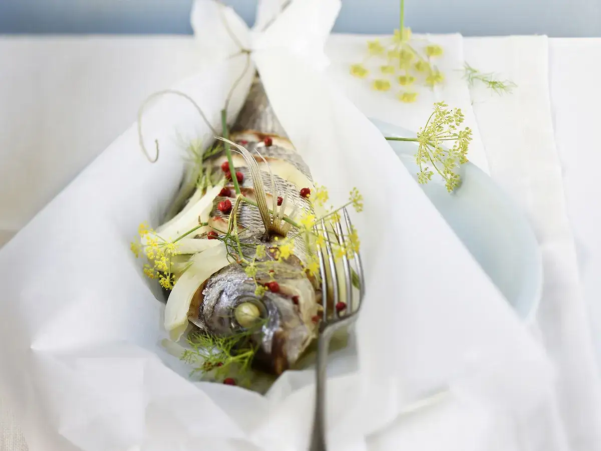 Dorade en papillote, un poisson entier cuit dans du papier blanc, garni de fenouil et de baies roses, prêt à être dégusté.
