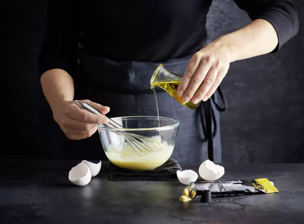 Préparation de mayonnaise maison : de l'huile est versée dans un bol contenant des œufs battus et un fouet. Des coquilles d'œufs et un tube de moutarde sont à proximité.