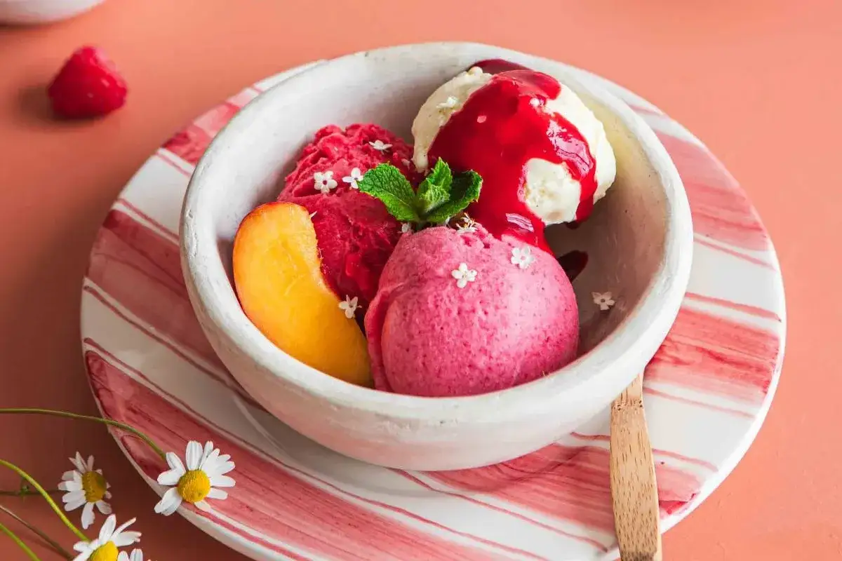 Un délice de pêche Melba : trois boules de sorbet aux fruits rouges et vanille, nappées de coulis, garnies de menthe et d'une tranche de pêche.