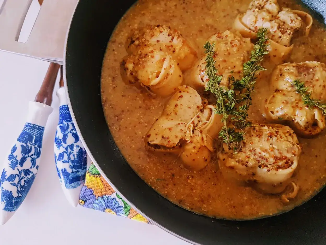 Cuisson paupiette de veau dans une sauce moutardée, garnie de thym frais. Des ustensiles décorés sont à côté.