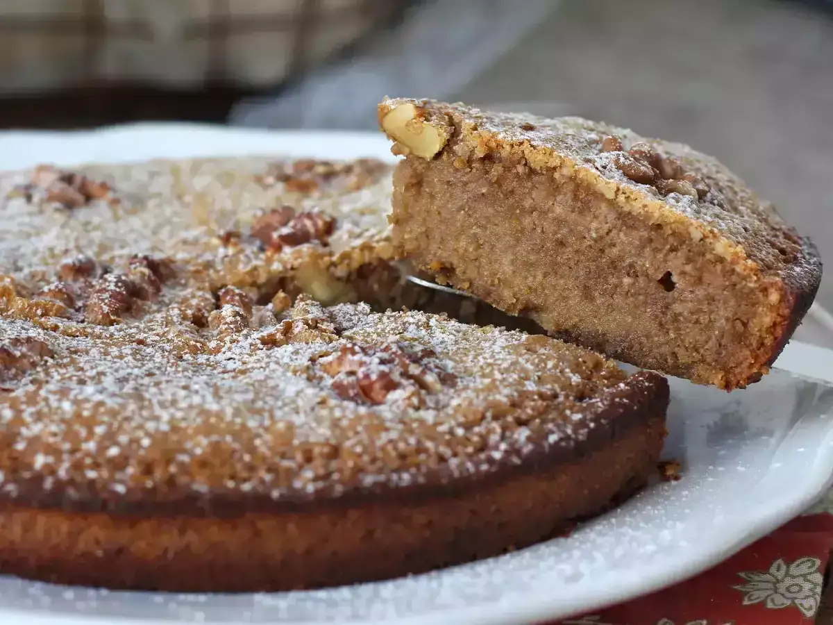 Un gâteau aux noix moelleux, saupoudré de sucre glace, est servi. Une part est soulevée, révélant sa texture.