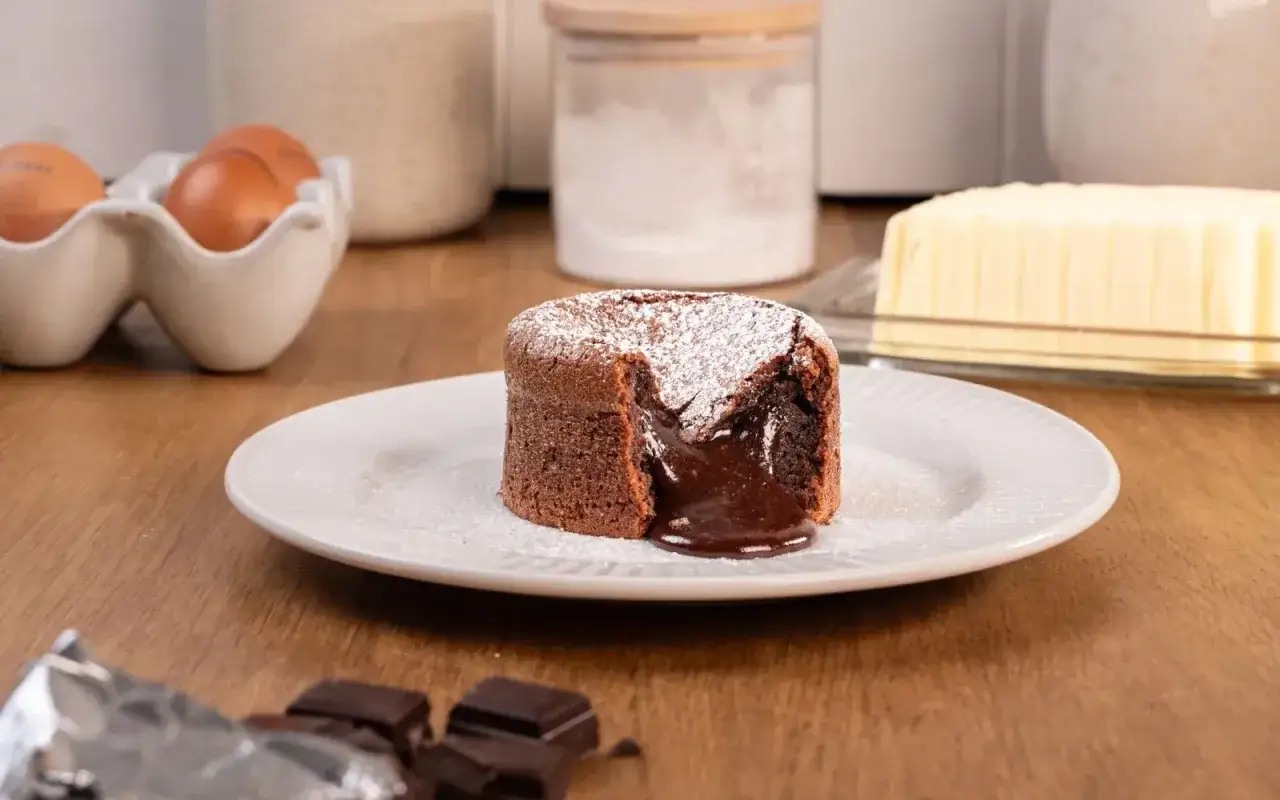 Un fondant au chocolat coulant, saupoudré de sucre glace, sur une assiette blanche. Des ingrédients de pâtisserie sont visibles en arrière-plan.