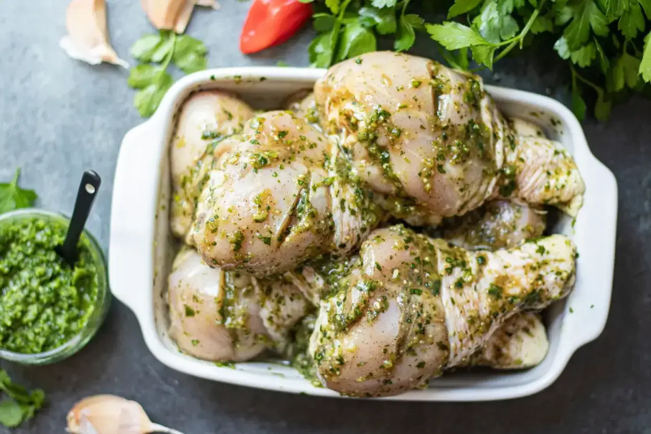Cuisses de poulet crues dans un plat blanc, recouvertes d'une marinade verte aux herbes. Prêtes pour la cuisson.
