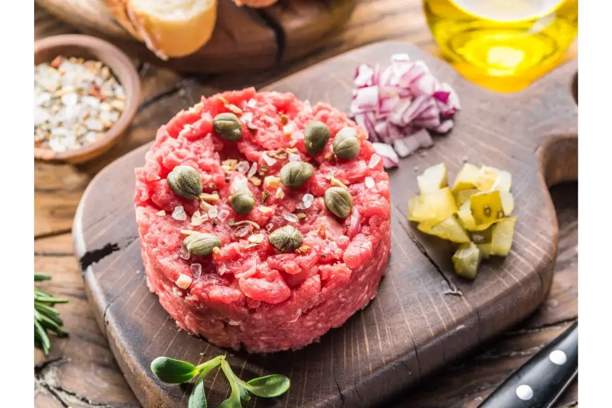 Un délicieux tartare de bœuf gastronomique, garni de câpres et de sel, accompagné d'oignons rouges et de cornichons sur une planche en bois.