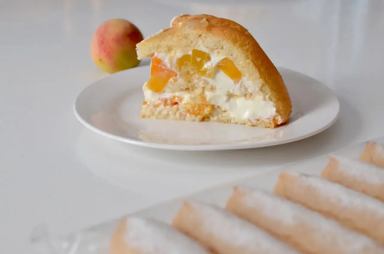 Une part de charlotte aux fruits et fromage blanc, garnie de morceaux de pêche, sur une assiette blanche. Un fruit entier est à côté.