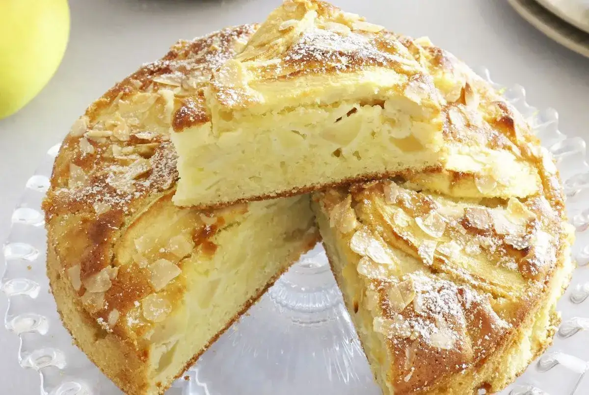 Un délicieux cake aux pommes, garni d'amandes effilées et saupoudré de sucre glace. Une part est déjà coupée, révélant la texture moelleuse.
