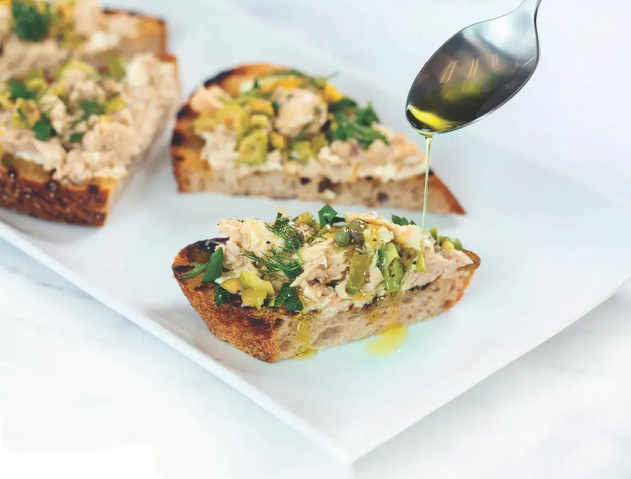 Tartines de pâté de thon rapide, garnies d'olives et d'herbes, arrosées d'huile d'olive.