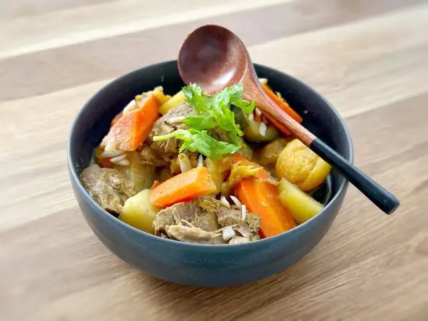 Un délicieux tajine agneau aux carottes et pommes de terre, garni de coriandre fraîche. Une cuillère en bois repose sur le plat.