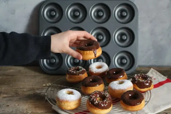 Une main tient un donut glacé au chocolat. Autour, plusieurs donuts, certains saupoudrés de sucre glace, d'autres de glaçage au chocolat et de vermicelles colorés, prêts à être dégustés. Une recette donuts professionnel réussie !