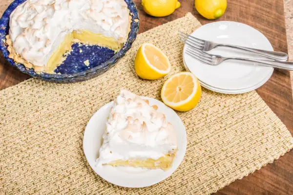 Une part de tarte citron meringuée sur une assiette, à côté d'une tarte entière et de citrons frais. Parfait pour une recette de tarte citron meringuée.
