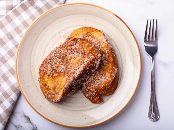 Deux tranches dorées de pain perdu au four, saupoudrées de sucre et de cannelle, sur une assiette avec une serviette à carreaux et une fourchette.