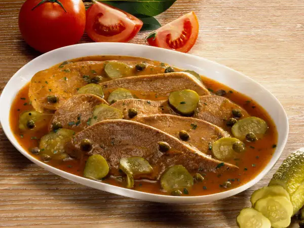 Langue de veau cuisson : fines tranches de langue de veau nappées d'une sauce savoureuse, garnies de cornichons et de câpres. Tomates en arrière-plan.