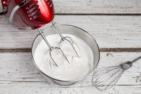 Un batteur rouge prépare une crème chantilly onctueuse dans un bol en inox. Un fouet repose à côté.