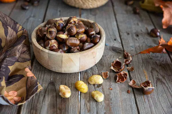 Châtaignes dorées et craquelées, fraîchement sorties de l'air fryer, dans un bol en bois. Quelques châtaignes décortiquées et leurs coquilles éparpillées sur la table en bois.