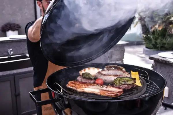 Un homme soulève le couvercle d'un barbecue fumant, révélant des saucisses, des steaks et du bacon. La cuisson lente permet d'attendrir la viande pour un repas savoureux.