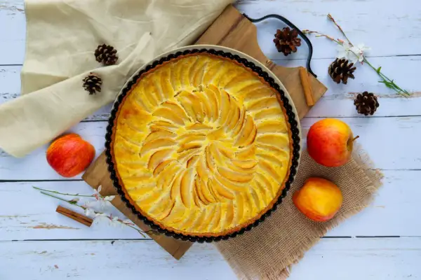 Une tarte aux pommes simple, dorée et appétissante, ornée de fines lamelles de fruits disposées en spirale. Des pommes fraîches, des pommes de pin et une branche de fleurs ajoutent une touche rustique.