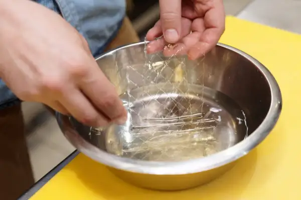 Mains trempant une masse gélatine en feuille dans un bol d'eau. La feuille de gélatine est transparente et a une texture réticulée.