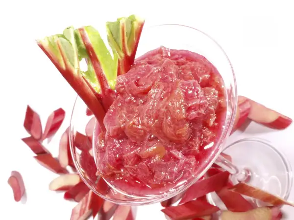 Un verre rempli de compote de rhubarbe, garni de tiges fraîches. Des morceaux de rhubarbe coupés entourent le verre.