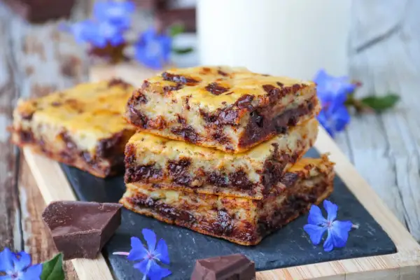 Gourmandises de cake banane pépite chocolat, empilées sur une ardoise, décorées de fleurs bleues et de morceaux de chocolat noir.