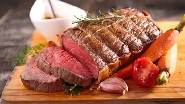 Un rôti de bœuf congelé au four, tranché, avec des carottes, une tomate, de l'ail et du romarin sur une planche en bois.