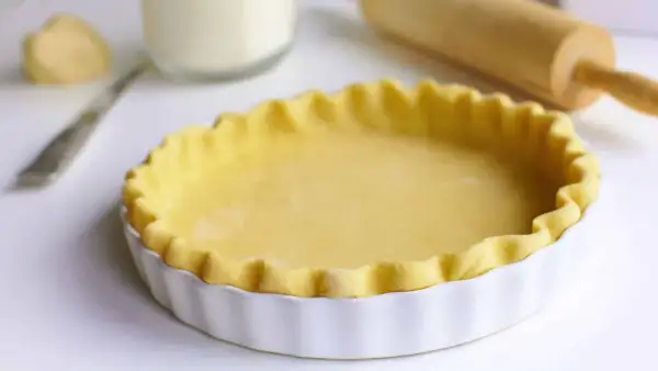 Pâte à tourte prête à être garnie, dans un plat blanc cannelé. Un rouleau à pâtisserie est visible en arrière-plan.