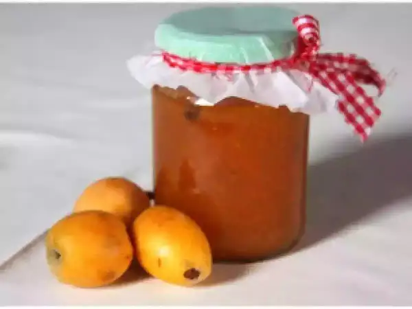 Un pot de confiture de nèfles, décoré d'un ruban à carreaux rouges et blancs, est posé à côté de trois fruits frais.