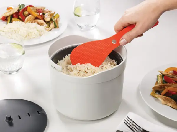 Une spatule orange sert du riz rond dans un cuiseur blanc. Le riz est prêt pour un repas savoureux.