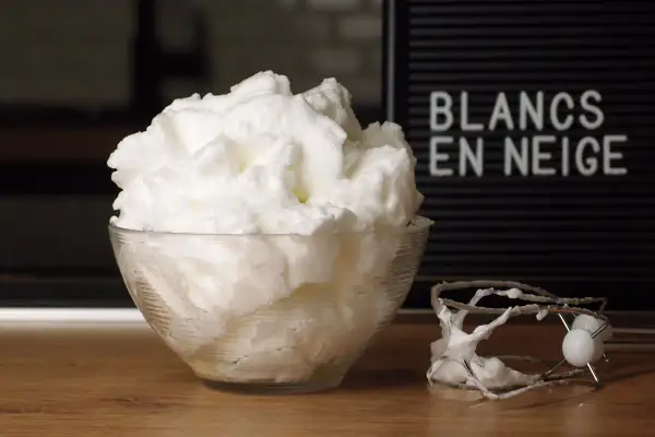 Un bol de blancs en neige aériens, comme un nuage, à côté d'un batteur. Le mot "BLANCS EN NEIGE" est visible sur un panneau.