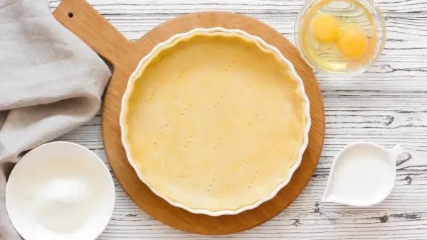 Une pâte à tarte crue est prête dans un plat blanc, entourée d'ingrédients pour différentes pâtes à tarte : œufs, lait, farine.